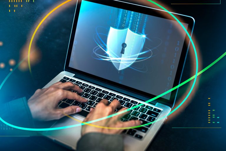 Hands typing on a laptop behind a glowing padlock shield, illustrating industrial IoT cybersecurity